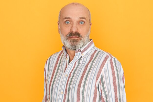 Astonished senior bald unshaven man wearing striped shirt opening eyes wide being caught by surprise, receiving sudden unexpected news posing isolated against yellow studio wall background