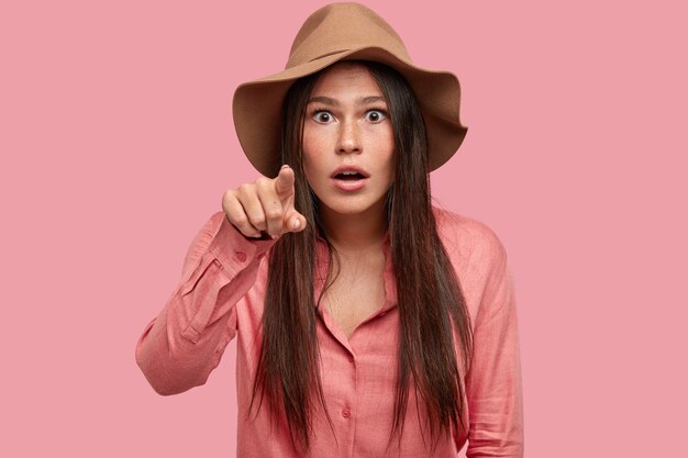 Astonished stupefied young freckled woman points with index finger at camera, opens mouth