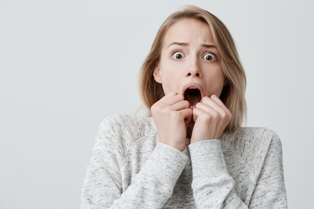 Astonished terrified attractive young blonde with dyed hair female in sweater having scared face expression, covering open mouth with hands