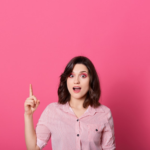 Astonished woman with good idea, pointing her finger up at copy space isolated over pink background, lady dresses casual attire, standing with opened mouth, looks surprised.