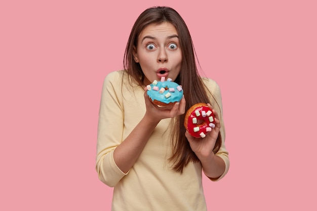 Foto gratuita stupito giovane carino donna europea birtes scintillante deliziosa ciambella, tiene gli occhi ben aperti, vestito con un maglione giallo, ha uno spuntino gustoso