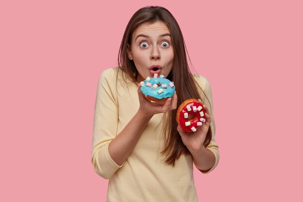 Astonished young cute European woman birtes sparkled delicious doughnut, keeps eyes wide opened, dressed in yellow sweater, has tasty snack