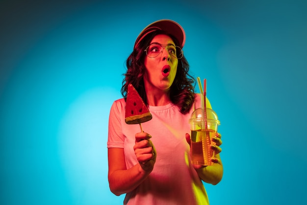 Astonished young woman standing with drink and sweets over trendy blue neon