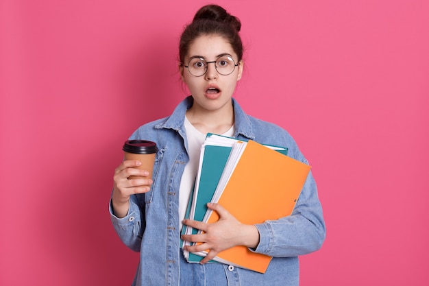 Astonished young woman with widely opened mouth, holding takeaway coffee and paper folder