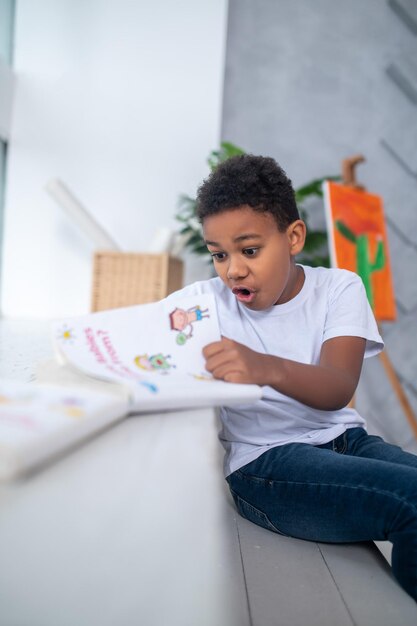 Astonishment. Dark-skinned school-age boy in white tshirt looking in surprise at book with open mouth sitting in bright room in daylight