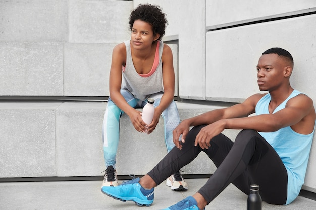 Free photo athlete female and male runner sit at stairs, being deep in thoughts, dressed in casual clothes, drink coffee from sport clothes, feel tired rest after jogging. people, motivation, fitness concept