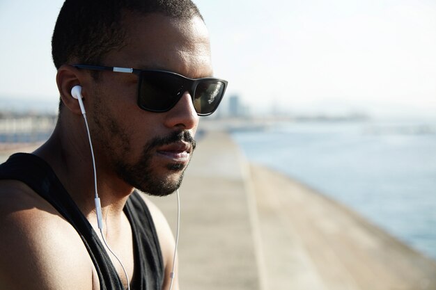 Athlete sitting on embankment on sunny summer day while resting after intensive training outdoors. Exhausted fit runner resting at seaside after workout. Sportsman listening to music with headphones