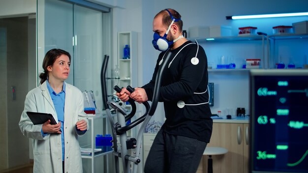 Спортсмен разговаривает с медицинским исследователем, бегающим на кросс-тренажере в спортивной научной лаборатории, измеряя максимальное значение VO2, частоту сердечных сокращений, психологическое сопротивление и мышечную выносливость