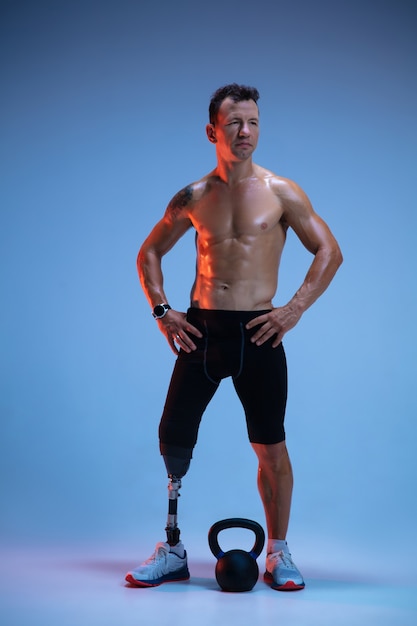 Athlete with disabilities or amputee isolated on blue studio background. Professional male sportsman with leg prosthesis training with weights in neon.