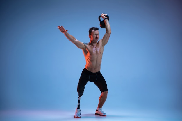 Athlete with disabilities or amputee isolated on blue studio background. Professional male sportsman with leg prosthesis training with weights in neon.