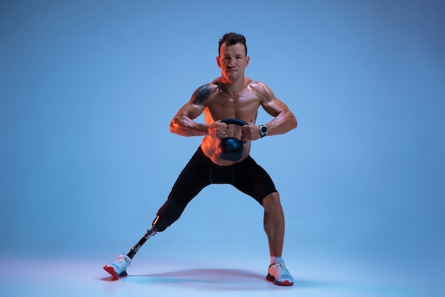 Athlete with disabilities or amputee isolated on blue studio background. professional male sportsman with leg prosthesis training with weights in neon.