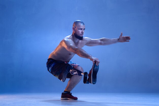 Athlete with disabilities or amputee isolated on blue  wall. Professional male sportsman with leg prosthesis training active in neon. Disabled sport and overcoming, wellness concept.