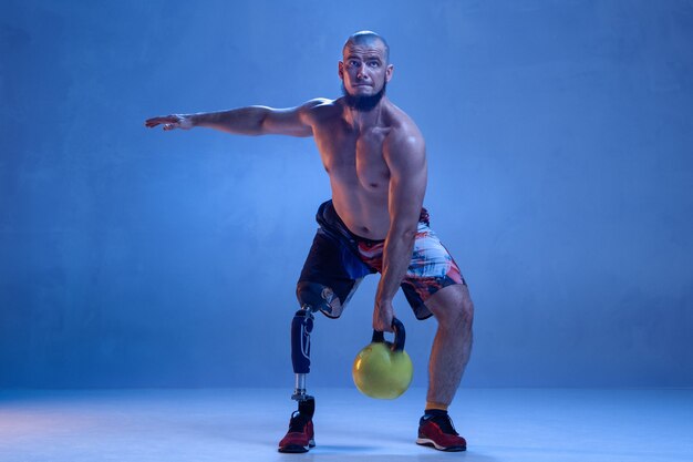 Athlete with disabilities or amputee isolated on blue  wall. Professional male sportsman with leg prosthesis training with weights in neon. Disabled sport and overcoming, wellness concept.