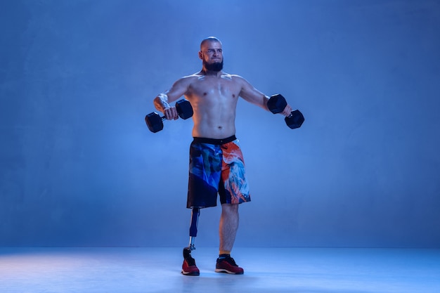 Free photo athlete with disabilities or amputee isolated on blue  wall. professional male sportsman with leg prosthesis training with weights in neon. disabled sport and overcoming, wellness concept.