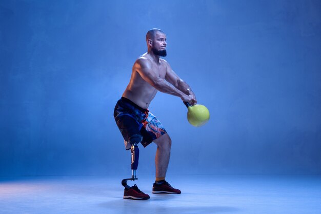 青い壁に孤立した障害のある運動選手または切断者。ネオンでウェイトトレーニングをしている義足のプロの男性スポーツマン。障害者スポーツと克服、ウェルネスの概念。