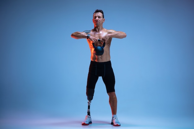 Спортсмен-инвалид или человек с ампутированной конечностью, изолированные на синей стене. Профессиональный спортсмен-мужчина с тренировкой протезирования ноги с гирями в неоне. Спорт для инвалидов и преодоление, концепция оздоровления.