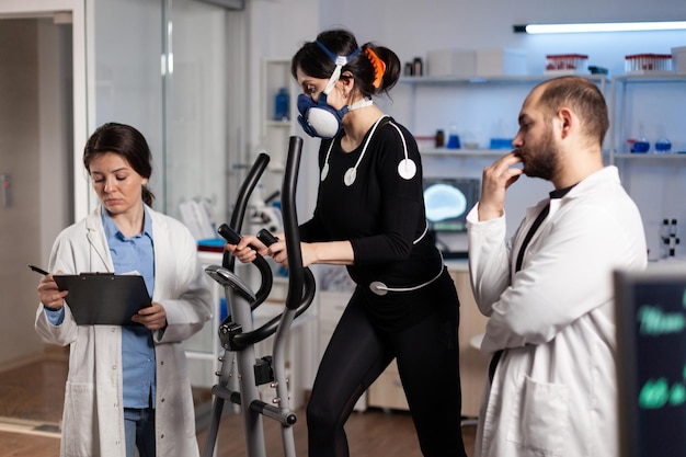 研究室で心拍数モニタリングegkデータを測定している研究者の医師が体の持久力を訓練している間、ジムのバイクルで走っているマスクを持ったアスリートの女性。その上に医療電極を持つスポーツウーマン