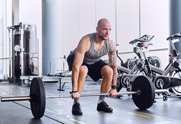 Athletic bearded shaved head male working out with barbell in a gym club.