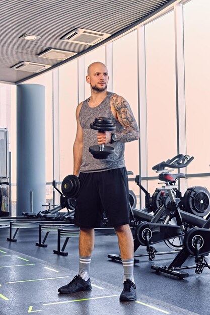 Athletic bearded shaved head male working out with dumbbells in a gym club.