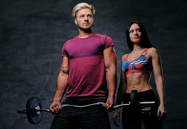 Free photo the athletic, blond male holds barbell weight and slim fitness female model over dark grey background.