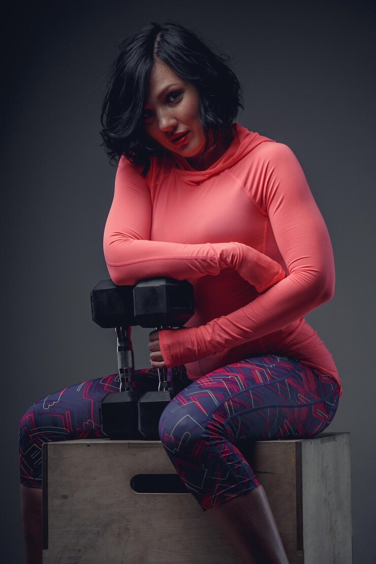 Free Photo athletic brunette woman in pink shirt sitting on wooden box and holding dumbell.