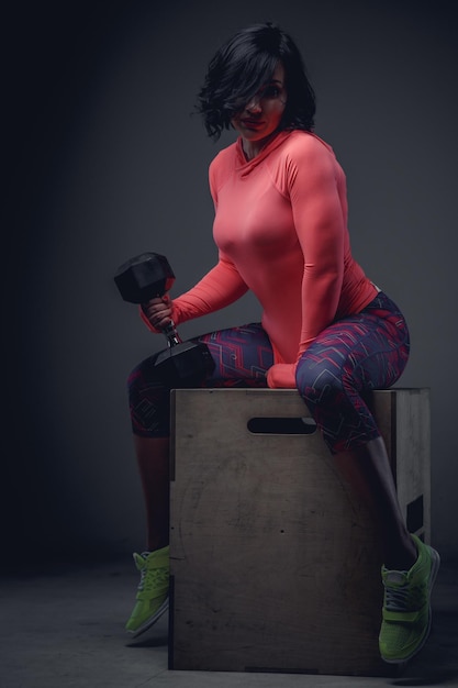 Athletic brunette woman in pink shirt sitting on wooden box and holding dumbell.