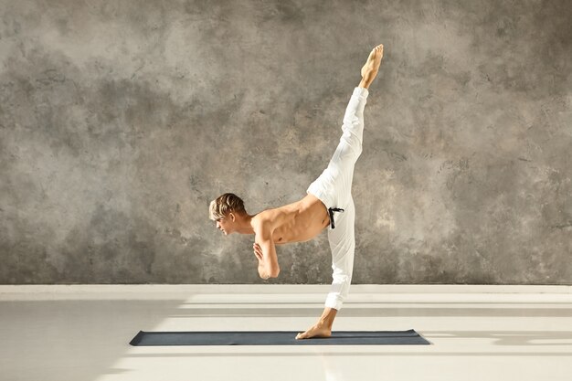 Athletic fit young male with muscular torso wearing no shirt practicing advanced yoga asana, standing with one leg on floor, training balance, concentration and coordination, bending forward