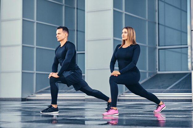 The athletic fitness couple is stretching over modern building background. Full body image.