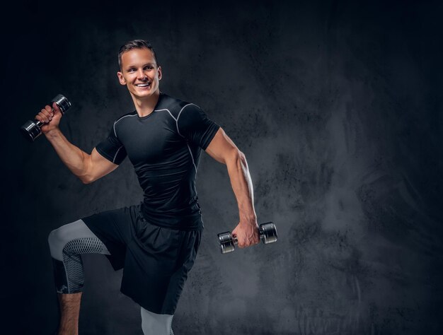 An athletic fitness male dressed in sportswear holds a set of dumbbells over grey background.