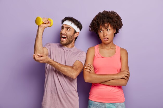 Athletic man lifts dumbbell, has weary workout, wears headband and t shirt, unhappy bored woman stands with arms folded