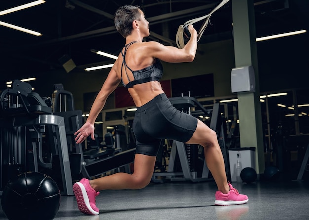 Athletic middle age female with short hair doing legs workouts with trx suspension strips in a gym club.