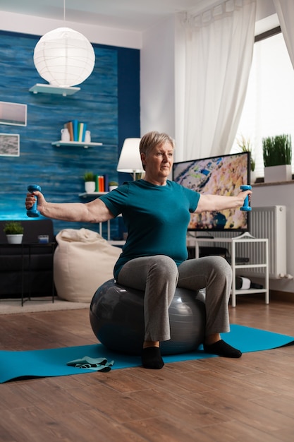 Athletic pensioner in sportswear watching online aerobic workout using tablet sitting on swiss ball