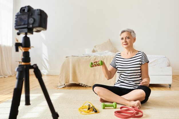 Athletic senior female fitness instructor with short gray hair exercising on floor with green dumbbells, recording video tutorial via camera on tripod. People, age and healthy active lifestyle