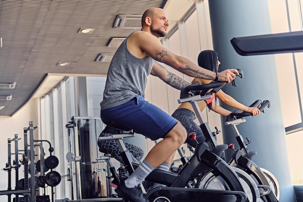 Спортивный мужчина с бритой головой и две стройные фитнес-женщины тренируются с велосипедом в тренажерном зале.