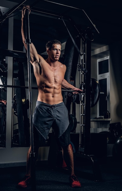 Athletic shirtless muscular male posing with barbell in a gym club.