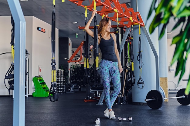 Спортивная стройная фитнес-модель в красочной спортивной одежде, позирующая рядом с полосками подвески trx, стоит в спортивном клубе.