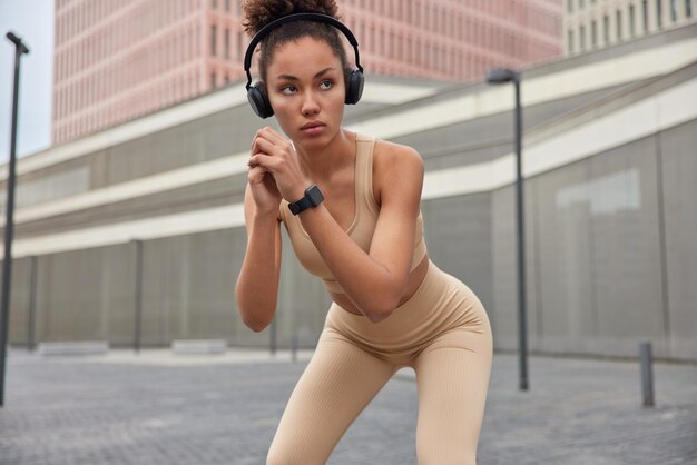 Athletic sporty woman squats outdoors dressed in sportswear makes cardio workout during morning keeps hands together concentrated serious away listens music via wireless headphones poses outside