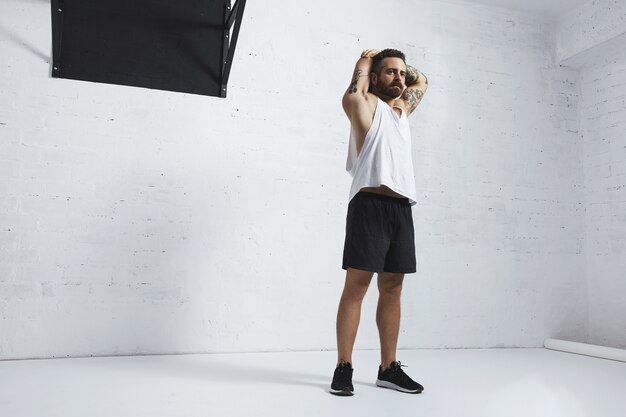 Athletic tattooed man in white blank tank t-shirt stretching his triceps on arms after workout, isolated on brick wall