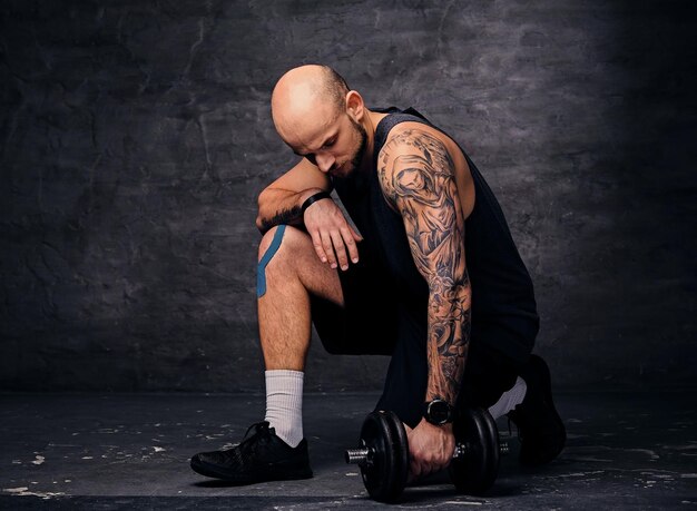 Athletic tattooed, shaved head sporty male standing in knee and holds dumbbell