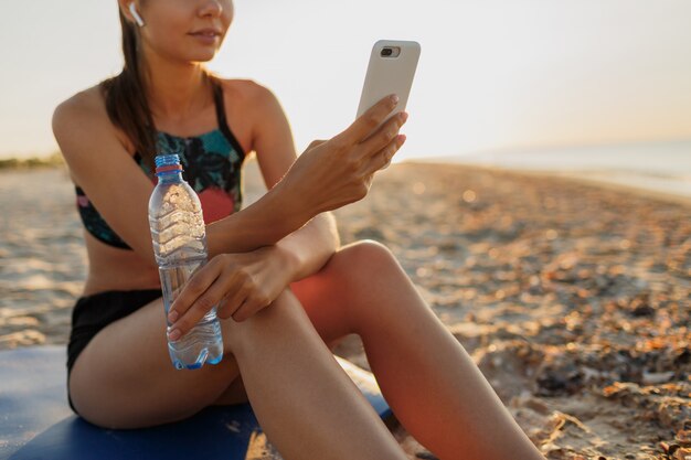 Спортивная (ый) женщина отдыхает после тренировки, слушает музыку, держит мобильный телефон и бутылку воды. Рано утром восход солнца на пляже.