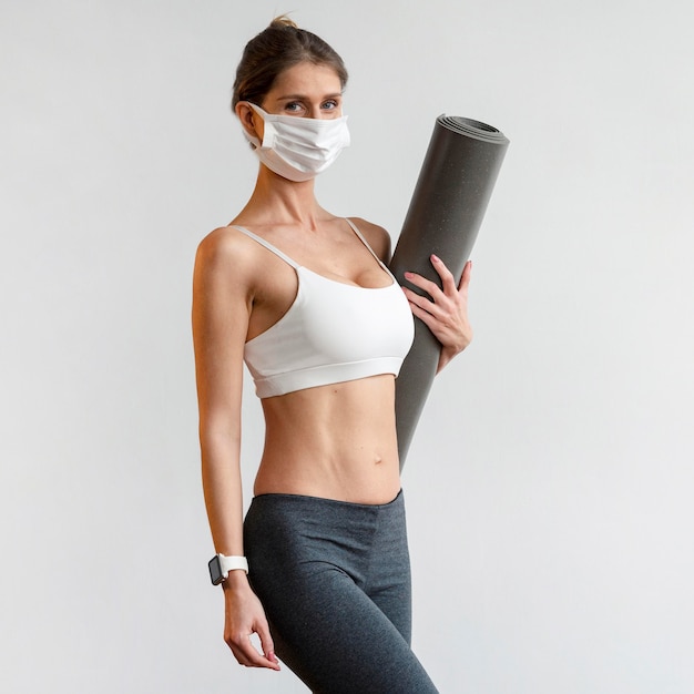 Athletic woman with medical mask holding yoga mat