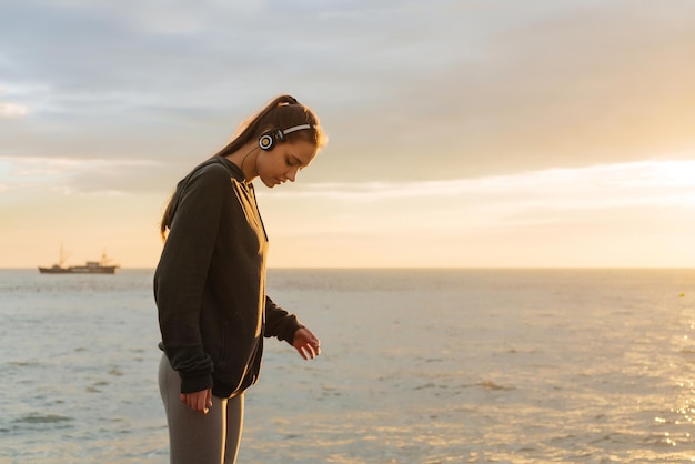 運動の若い女の子は、ヘッドフォンで音楽を聴きながら、日没時に海の景色を楽しんでいます Premium写真