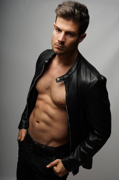Athletic young Hispanic male model wearing a black leather jacket and posing against a gray wall
