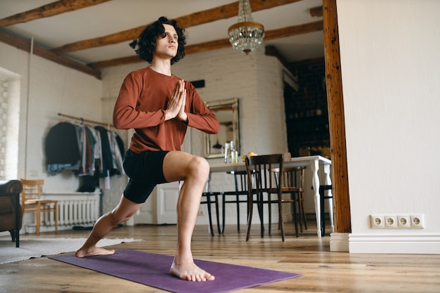 Athletic young male yogi practicing yoga indoors, standing barefooted on mat, holding hands in namaste, doing sun salutation sequence in the morning.