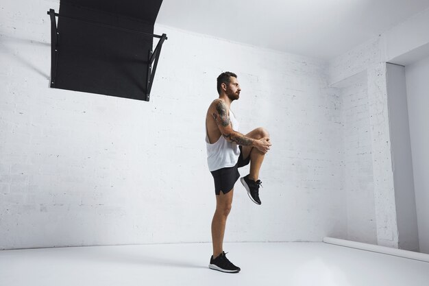 Athletic young man doing knee raises, stretching his legs, looking right side, isolated on white brick wall next to pull bar