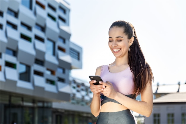 La giovane donna atletica sta sorridendo e sta guardando il telefono cellulare in sue mani