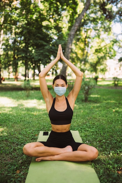 医療用防毒マスクを着用した運動の若い女性、午前中に公園でヨガをしている、ヨガマットでの女性のトレーニング