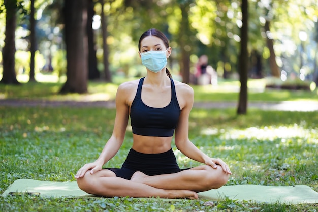 医療用保護マスクを着用したアスレチック若い女性、午前中に公園でヨガをしている、ヨガマットでの女性のトレーニング