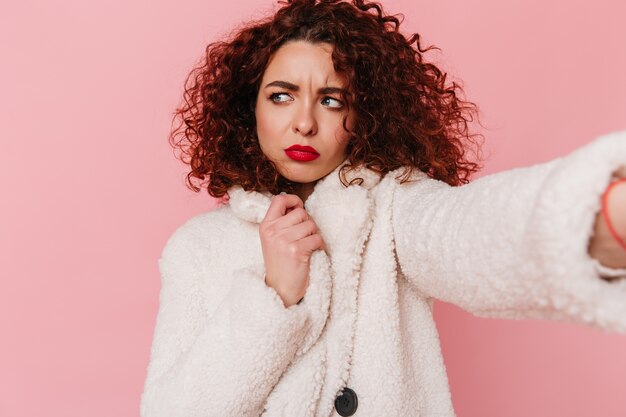 Atractive curly girl with red lips dressed in white wool coat makes selfie on pink isolated space.