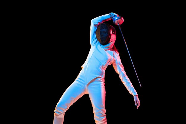 Attacking. Teen girl in fencing costume with sword in hand isolated on black background, neon light. Young model practicing and training in motion, action. Copyspace. Sport, youth, healthy lifestyle.